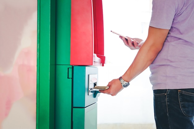 Zdjęcie młody człowiek gospodarstwa smartphone i za pomocą bankomatu automated teller machine