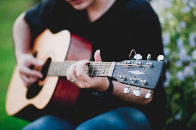 młody człowiek gra na gitarze, siedząc w naturalnym ogrodzie