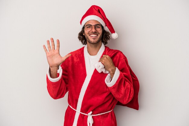 Młody człowiek kaukaski przebrany za Świętego Mikołaja na białym tle na szarym tle uśmiechający się wesoły Wyświetlono numer pięć palcami.