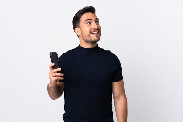 Młody człowiek kaukaski przy użyciu telefonu komórkowego na białym tle na białej ścianie patrząc w górę podczas uśmiechu