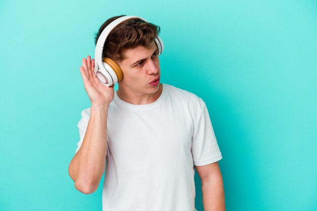 Młody człowiek kaukaski, słuchanie muzyki w słuchawkach na białym tle na niebieskiej ścianie, próbując słuchać plotek.