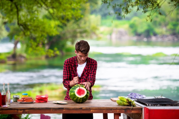 młody człowiek kroi soczystego arbuza podczas francuskiej kolacji na świeżym powietrzu w pobliżu rzeki w piękny letni wieczór na łonie natury