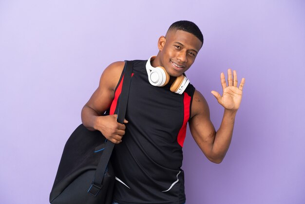 Młody człowiek Łacińskiej sportu z torbą sportową na białym tle na fioletowy salutowanie ręką z happy wypowiedzi