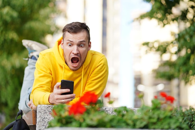 Młody człowiek leżał na brzuchu na ławce z telefonem w rękach i w szoku, co zobaczył na ekranie smartfona