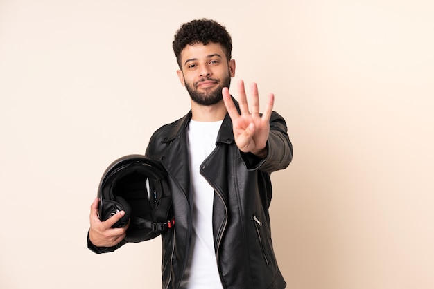 Młody człowiek Maroka w kasku motocyklowym na białym tle na beżowej ścianie szczęśliwy i liczy cztery palcami