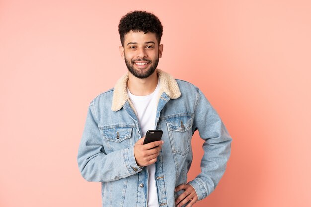 Młody człowiek Maroka za pomocą telefonu komórkowego na białym tle na różowej ścianie, pozowanie z rękami na biodrze i uśmiechnięty