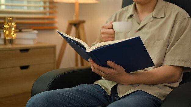 Młody człowiek pijący kawę i czytający nową powieść siedząc na wygodnym fotelu w domu