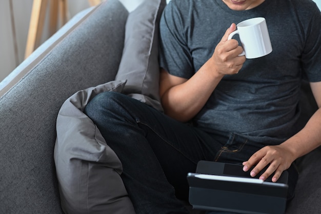 Młody człowiek pije kawę i czyta e-mail na tablecie komputerowym.