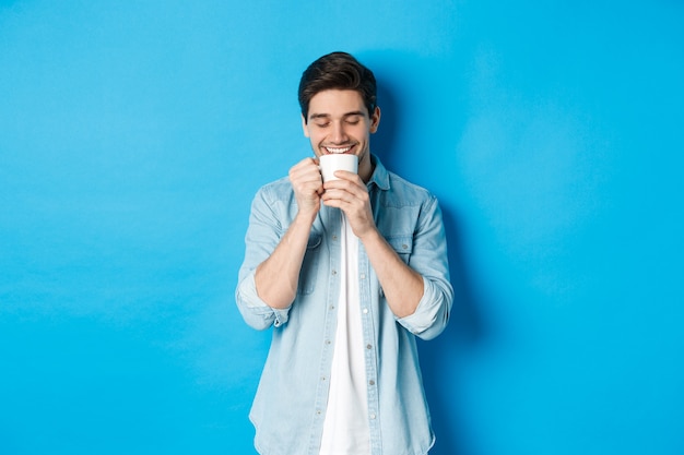 Młody człowiek pije kawę z radosną buźką, stojąc na niebieskiej ścianie