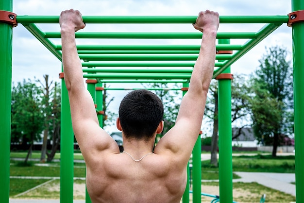 Młody człowiek podciąga się na poziomym pasku na zewnątrz na boisku sportowym. Street Workout.