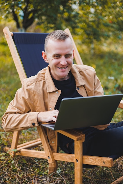 Młody człowiek pracuje na laptopie na zewnątrz Młody freelancer odpoczywa w lesie Praca zdalna aktywny wypoczynek latem Turystyka ludzie koncepcja mężczyzna siedzi na krześle na zewnątrz