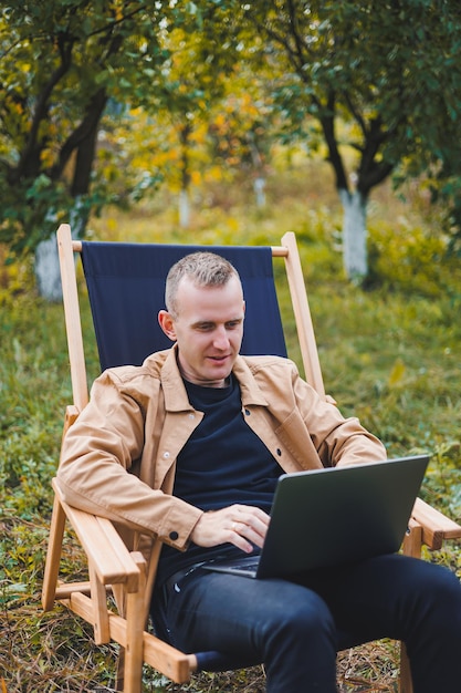 Młody człowiek pracuje na laptopie na zewnątrz Młody freelancer odpoczywa w lesie Praca zdalna aktywny wypoczynek latem Turystyka ludzie koncepcja mężczyzna siedzi na krześle na zewnątrz