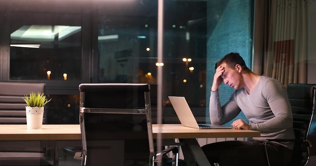 Młody człowiek pracuje na laptopie w nocy w ciemnym biurze. Projektant pracuje w późniejszym czasie.