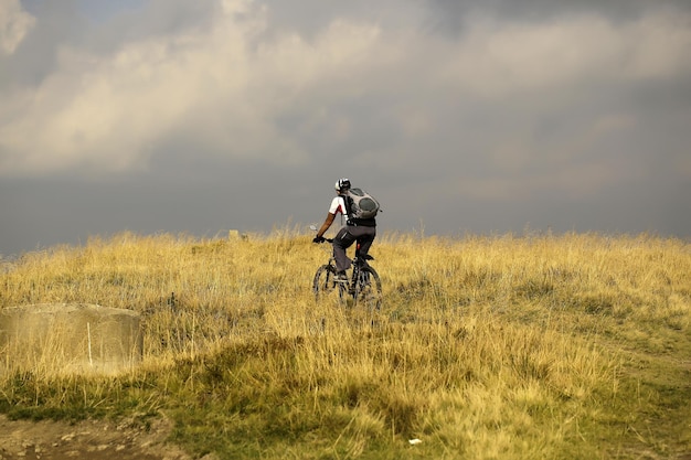 Młody człowiek rowerzysta z plecakiem jeździ na rowerze górskim na polu trawy na szarym niebie