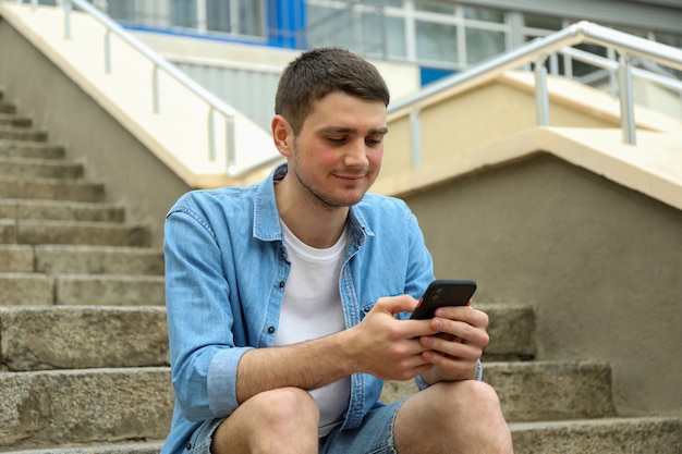 Młody człowiek siedzi na schodach i patrząc w telefonie