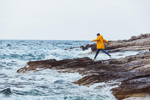 Młody człowiek skaczący na skalistym wybrzeżu w żółtym płaszczu przeciwdeszczowym w przestrzeni kopii pogody sztormowej
