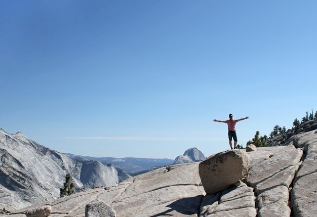 Młody człowiek stojący na skale z podniesionymi rękami przed panoramą pasma górskiego Yosemite