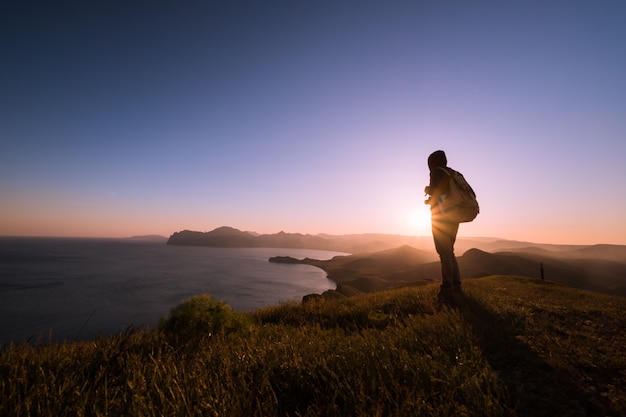 Młody człowiek stojący z plecakiem. Wycieczkowicz na kamieniu nad brzegiem morza w kolorowe niebo słońca. Piękny krajobraz ze sportowym mężczyzną kołysze morze i chmury o zachodzie słońca. Sportowy styl życia