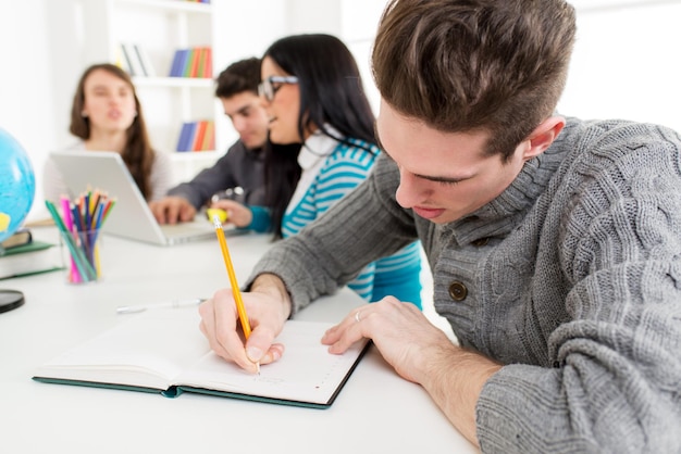 Młody człowiek student siedzi i pisze na pierwszym planie. Za nim siedzi szczęśliwa grupa przyjaciół i patrzy na laptopa.