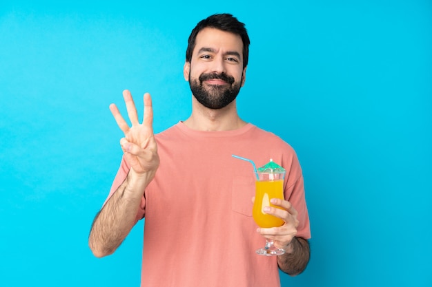 Młody człowiek szczęśliwy trzyma koktajl nad odosobnioną błękit ścianą szczęśliwą i liczy trzy z palcami
