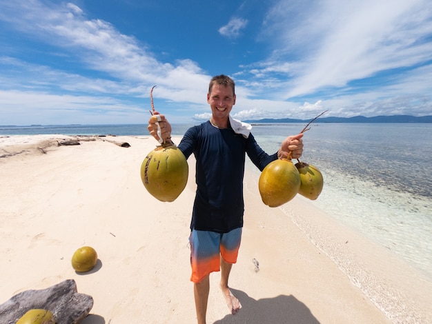Młody człowiek trzyma kokosy na piaszczystej plaży z wodą i niebem na tle. Koncepcja przygody i podróży.