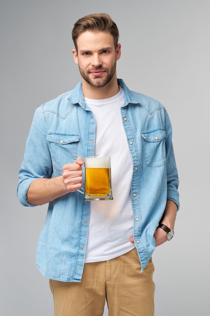 Młody człowiek trzyma na sobie dżinsy koszuli trzymając szklankę piwa stojąc na szarej ścianie.