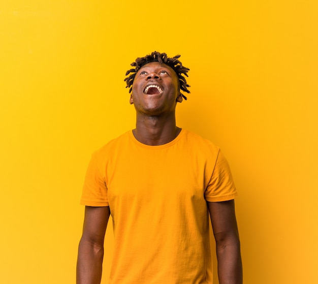 Młody człowiek ubrany w rastas na żółtej ścianie zrelaksowany i szczęśliwy śmiejąc się, szyja rozciągnięta pokazując zęby
