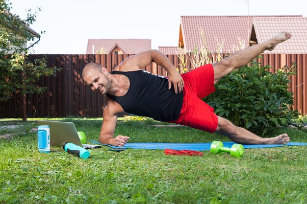 Młody człowiek uprawia sport w parku. Sportowiec o blond włosach robi deskę, ogląda film i studiuje z laptopa na dywanie na podwórku w letni dzień