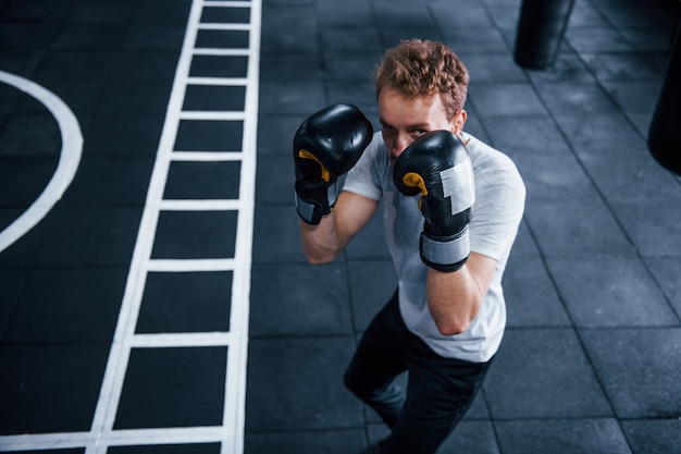 Młody człowiek w białej koszuli i bokserskich rękawicach ochronnych robi ćwiczenia w siłowni.