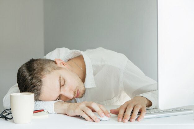 Zdjęcie młody człowiek w białej koszuli leżącego na pulpicie z komputerem w biurze. pracownik na szarym tle. odpoczywający człowiek
