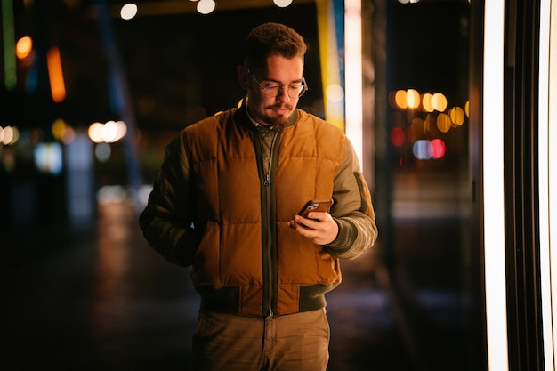 Zdjęcie młody człowiek w nocy spaceruje i patrzy na swój smartfon z niewyraźnym tłem