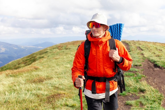 Młody człowiek w pomarańczowej kurtce i okularach chodzi po górach z kijem do turystyki.