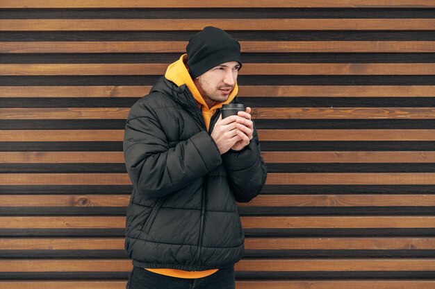 Młody człowiek w zimowych ubraniach stojący z kawą, aby wyjść na ulicę przed drewnianą ścianą