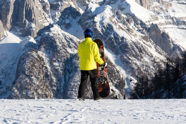 Młody człowiek w żółtej kurtce chodzi po stoku narciarskim z snowboardem