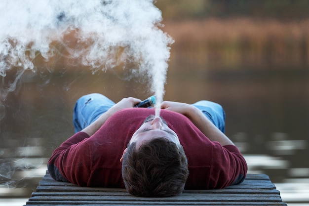 Zdjęcie młody człowiek waper leży na moście w jesienny dzień i pali e-papierosa