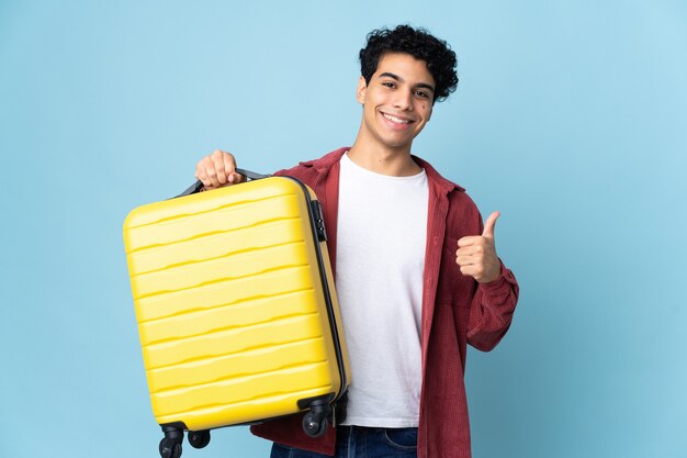 Młody człowiek Wenezueli na białym tle na niebieskim tle w wakacje z walizką podróżną iz kciukiem do góry