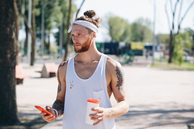 Młody człowiek z białą opaską na głowie ubrany w białą koszulkę za pomocą telefonu komórkowego i trzyma plastikowy kubek w ręku na boisku sportowym na zewnątrz w słoneczny dzień.