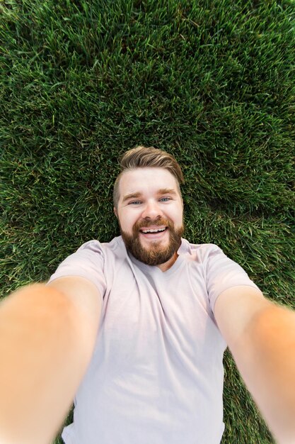 Młody człowiek z brodą i modą fryzurą, leżąc na trawie, biorąc selfie, trzymając inteligentny telefon lub tablet i patrząc na kamerę