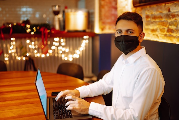 Młody człowiek z maską ochronną pracuje na laptopie w domu podczas ferii zimowych.