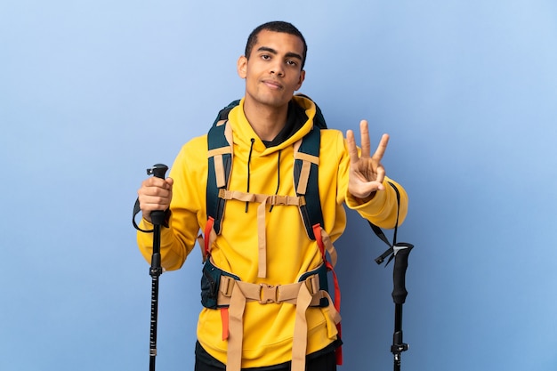 Młody człowiek z plecakiem i kijkami trekkingowymi nad izolowaną ścianą szczęśliwy i liczy trzy palcami