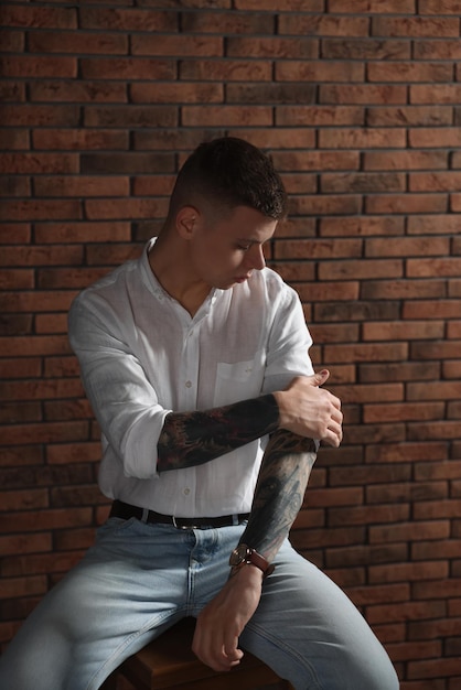 Młody człowiek z tatuażami siedzi na stołku w pobliżu ściany z cegły