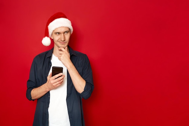 Młody człowiek z telefonem w dłoni uśmiecha się chytrze, myśli o nowym pomyśle, gratulacje, świąteczny santa hat