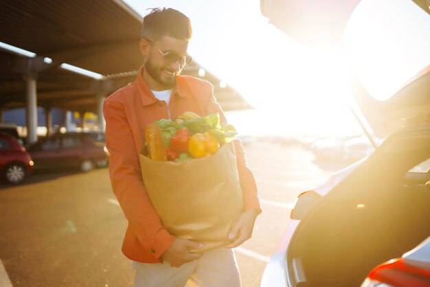 Młody człowiek z torbą na zakupy pełną warzyw w pobliżu samochodu Przystojny mężczyzna po zakupach