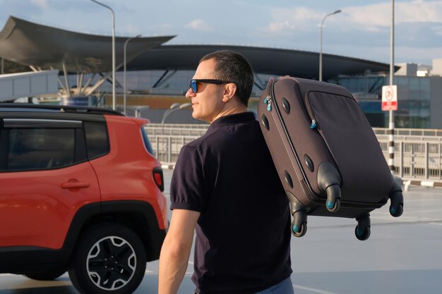 Młody człowiek z walizką w ręku w pobliżu samochodu na parkingu w słoneczny dzień