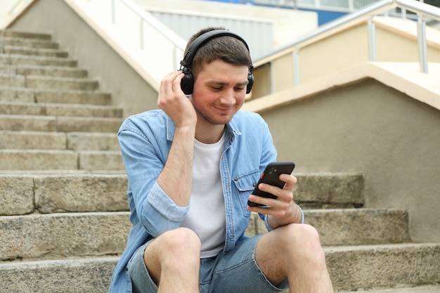 Młody człowiek ze słuchawkami siedzi na schodach i patrzy na swój telefon