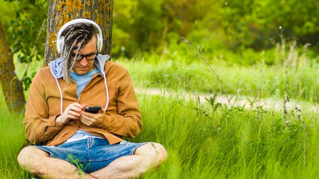 Młody człowiek zrelaksowany w słuchawkach okulary siedzi w pobliżu drzewa w zielonej trawie, słuchając muzyki na telefonie komórkowym Beztroski facet relaksujący się w naturze, słuchając piosenek na gadżecie w okresie letnim