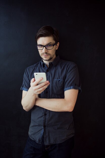 Młody dorosły facet w okularach używa smartfona. Biznesowy portret na textured czerni