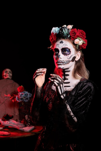 Młody dorosły noszący sztukę ciała dios de los muertos, aby wyglądać jak tradycyjna czaszka cartina w studio, mając koronę z kwiatów i kostium świętego mikołaja. Meksykańska tradycja świąteczna w dzień halloween.