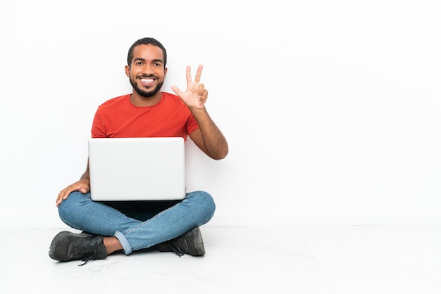 Młody Ekwadorczyk Z Laptopem Siedzący Na Podłodze Na Białym Tle Szczęśliwy I Liczący Trzy Palcami