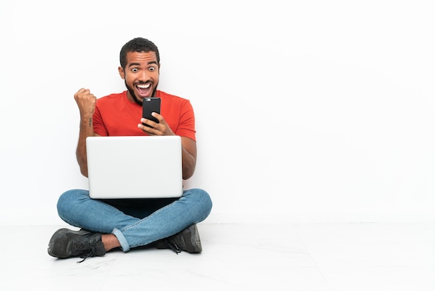 Młody Ekwadorczyk z laptopem siedzący na podłodze na białym tle zaskoczony i wysyłający wiadomość